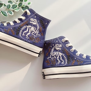 Op de afbeelding: Een paar hoge canvas sneakers in een diepblauwe kleur. De sneakers hebben een witte tussenzool en veters, met een gedetailleerd geborduurd ontwerp van een dinosaurus skelet en botanische elementen in wit en bruin.
