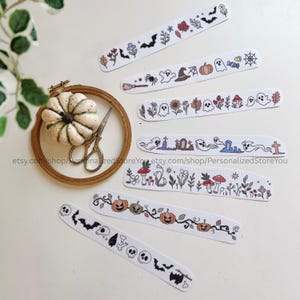 Op de afbeelding: Zeven lange, smalle papieren stroken met kleurrijke, handgetekende illustraties van Halloween-thema's, waaronder geesten, pompoenen, vleermuizen, schedels, paddenstoelen, bloemen en grafstenen, naast een houten borduurring met een pompoen.