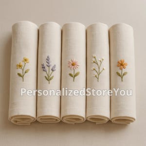 May include: Five rolled, off-white linen napkins, each embroidered with a different flower: yellow, lavender, pink, white, and orange. The text "PersonalizedStoreYou" is printed on the napkins.
