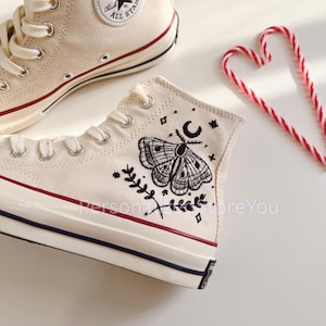 Può includere: Scarpe da ginnastica alte color crema con un design a falena nera e celeste. Le scarpe hanno lacci bianchi, una striscia rossa e una striscia nera e blu sulla suola. Il testo "Personal Store You" è visibile.