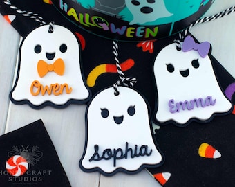 Könnte beinhalten: Weiße, geisterförmige Halloween-Namensschilder mit schwarzem Rand, mit Namen wie "Owen", "Emma" und "Sophia". Jeder Geist hat ein einzigartiges Accessoire: eine orangefarbene Fliege oder eine lila Schleife. Die Anhänger hängen an schwarz-weißem Bindfaden.