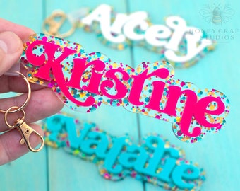 May include: Three colorful acrylic keychains with names "Kristine", "Natalie", and "Ace" written in a whimsical font. The keychains are filled with colorful glitter and have a gold metal key ring.