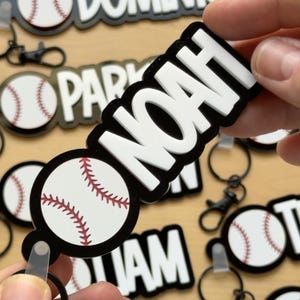 Könnte beinhalten: Baseball-Schlüsselanhänger mit dem Namen "NOAH" in weißen, fetten Buchstaben auf schwarzem Hintergrund. Der Schlüsselanhänger zeigt auch eine Baseball-Grafik mit roten Nähten. Weitere Schlüsselanhänger mit Namen und Baseball-Grafiken sind sichtbar.