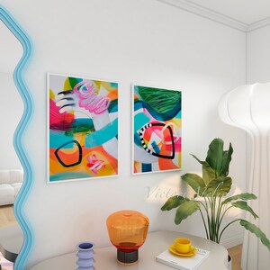 Puede incluir: Dos pinturas abstractas con colores brillantes y formas geom&eacute;tricas cuelgan de una pared blanca. Una l&aacute;mpara blanca con una pantalla redonda y grande se encuentra sobre una mesa con un libro, una taza y un platillo, y un jarr&oacute;n.