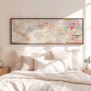 May include: Abstract art print in a rectangular black frame. The artwork features a mix of colors, including pink, beige, and light blue, with various shapes and textures. The print hangs above a bed with white and beige bedding.