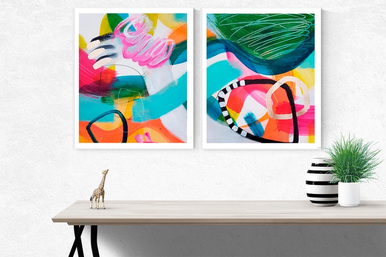 Puede incluir: Dos pinturas abstractas en marcos blancos, una con formas rosas, naranjas, amarillas, azules y verdes, la otra con formas turquesa, verdes, negras, blancas y doradas.