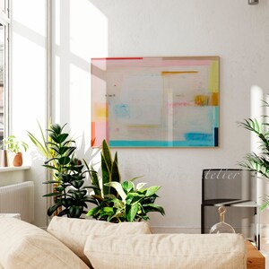 Puede incluir: Un interior de sala de estar con una gran pintura abstracta en un marco de madera clara en una pared blanca. La pintura presenta un fondo azul claro con formas geom&eacute;tricas rosas, amarillas y azules. Hay varias plantas de interior en macetas en el suelo, incluyendo una planta serpiente, una higuera de hoja de viol&iacute;n y una lirio de la paz. Un sof&aacute; beige con dos almohadas est&aacute; en primer plano.