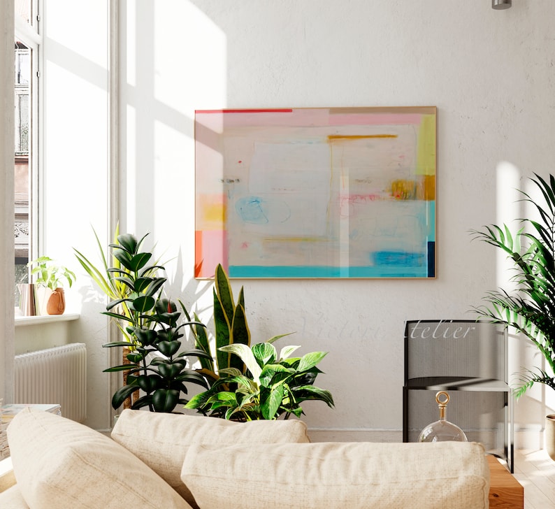 Puede incluir: Un interior de sala de estar con una gran pintura abstracta en un marco de madera clara en una pared blanca. La pintura presenta un fondo azul claro con formas geom&eacute;tricas rosas, amarillas y azules. Hay varias plantas de interior en macetas en el suelo, incluyendo una planta serpiente, una higuera de hoja de viol&iacute;n y una lirio de la paz. Un sof&aacute; beige con dos almohadas est&aacute; en primer plano.
