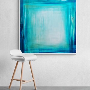 Pode incluir: Impress&atilde;o de arte abstrata com uma composi&ccedil;&atilde;o quadrada em tons de verde-azulado e turquesa, emoldurada em branco. Uma moderna banqueta branca com pernas de madeira est&aacute; posicionada em frente &agrave; obra de arte, criando uma est&eacute;tica minimalista.