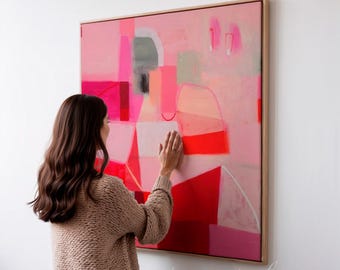 May include: Abstract artwork in a wooden frame. The painting displays a blend of pink, red, and grey geometric forms. A person's hand is touching the canvas. The artwork is mounted on a white wall.