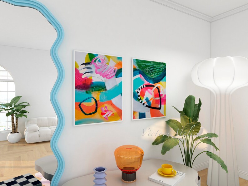 Puede incluir: Dos pinturas abstractas con colores brillantes y formas geom&eacute;tricas cuelgan de una pared blanca. Una l&aacute;mpara blanca con una pantalla redonda y grande se encuentra sobre una mesa con un libro, una taza y un platillo, y un jarr&oacute;n.