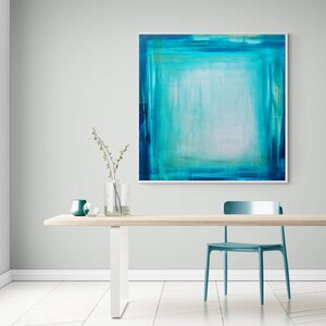 Pode incluir: Uma pintura abstrata em tons de verde-azulado e turquesa pendurada em uma parede cinza claro. A obra de arte, emoldurada em branco, apresenta uma &aacute;rea central clara cercada por tons mais escuros. Uma mesa de madeira com uma cadeira e vasos verde-azulado completam a cena.