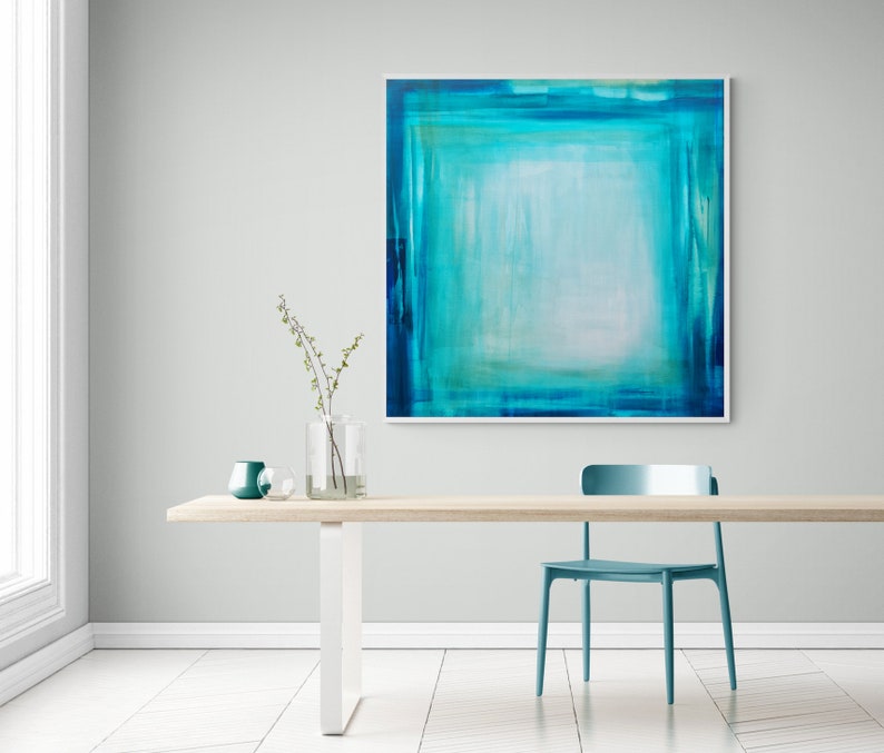 Pode incluir: Uma pintura abstrata em tons de verde-azulado e turquesa pendurada em uma parede cinza claro. A obra de arte, emoldurada em branco, apresenta uma &aacute;rea central clara cercada por tons mais escuros. Uma mesa de madeira com uma cadeira e vasos verde-azulado completam a cena.