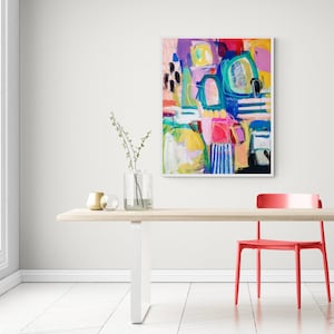 May include: Abstract painting featuring vibrant colours, including pink, blue, green, yellow, and white. The painting is framed in a white frame and hangs on a white wall above a wooden table with a red chair.