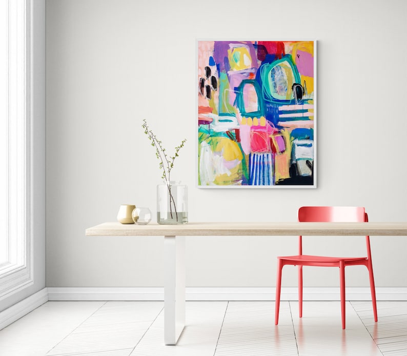 May include: Abstract painting featuring vibrant colours, including pink, blue, green, yellow, and white. The painting is framed in a white frame and hangs on a white wall above a wooden table with a red chair.