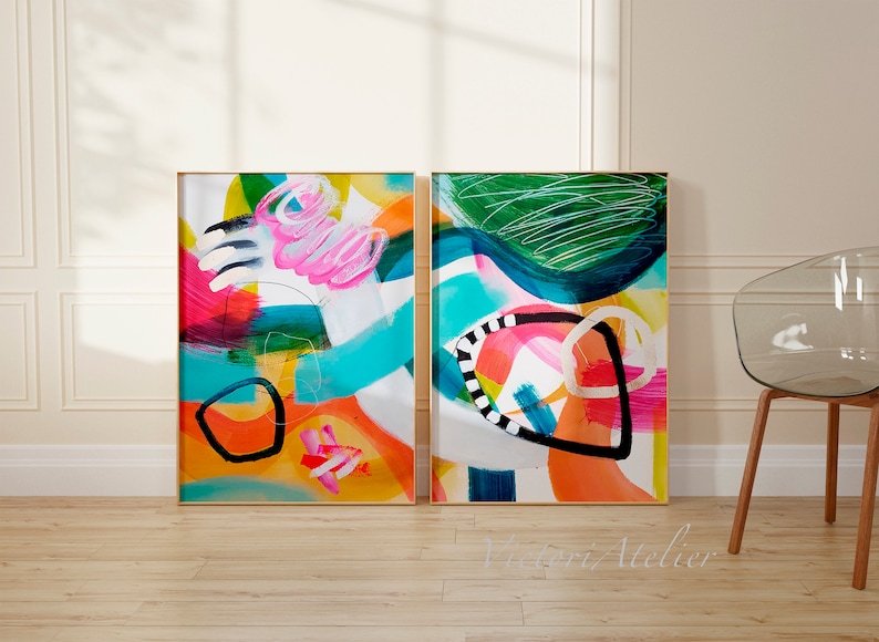 Puede incluir: Dos pinturas abstractas en marcos dorados con pinceladas audaces y coloridas. Las pinturas presentan una mezcla de colores brillantes, incluyendo rosa, naranja, amarillo, verde, azul y negro. Las pinturas est&aacute;n colocadas contra una pared blanca con un suelo de madera.