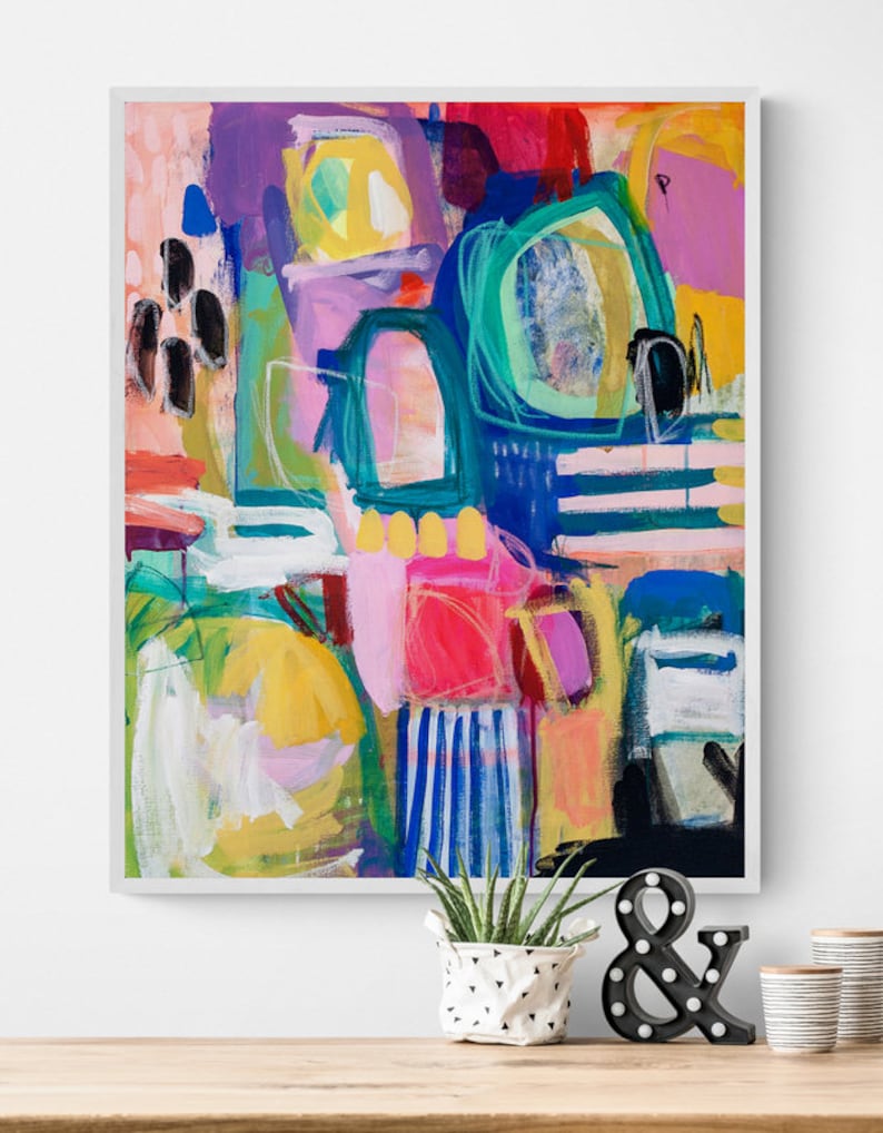 May include: Abstract painting with bold strokes of pink, yellow, blue, green, and black. The painting is framed in a white frame and is hanging on a white wall.