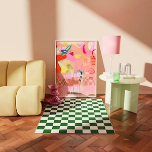 Op de afbeelding: Een gele bank met een groen-wit geblokt tapijt ervoor. Een ingelijst abstract schilderij met roze, gele en rode kleuren hangt aan de muur. Een roze lamp met een witte voet staat op een lichtgroene bijzettafel.