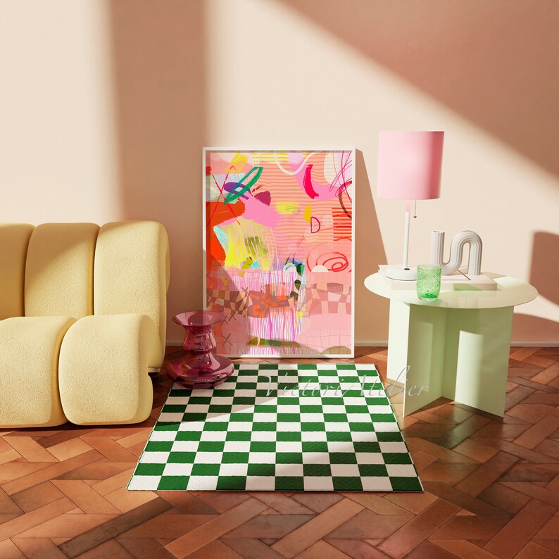 Op de afbeelding: Een gele bank met een groen-wit geblokt tapijt ervoor. Een ingelijst abstract schilderij met roze, gele en rode kleuren hangt aan de muur. Een roze lamp met een witte voet staat op een lichtgroene bijzettafel.