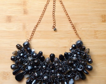 May include: A black beaded statement necklace with a copper chain. The necklace is made of various sizes and shapes of black beads, including round, oval, and teardrop shapes.