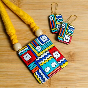 Puede incluir: Un conjunto de collar y pendientes coloridos. El collar presenta un colgante rectangular grande con un dise&ntilde;o geom&eacute;trico en rojo, azul, amarillo y blanco. Los pendientes tienen un dise&ntilde;o similar. El cord&oacute;n del collar es amarillo.