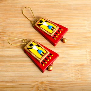 May include: A pair of red and yellow clay earrings with a gold trim. Each earring features a stylized figure in a white shirt and blue skirt.