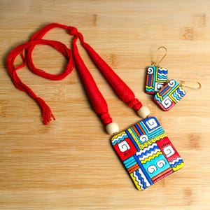 Puede incluir: Un conjunto de collar y pendientes hechos a mano con un colgante rectangular y pendientes a juego. La joyer&iacute;a tiene un dise&ntilde;o geom&eacute;trico rojo, azul, amarillo y blanco. El collar tiene un cord&oacute;n rojo y cuentas de madera.