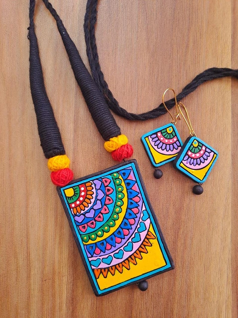 Puede incluir: Un conjunto de collar y pendientes coloridos con un colgante rectangular y pendientes a juego. El colgante y los pendientes presentan un dise&ntilde;o geom&eacute;trico con fondo amarillo y detalles azules, verdes, rosas y naranjas.