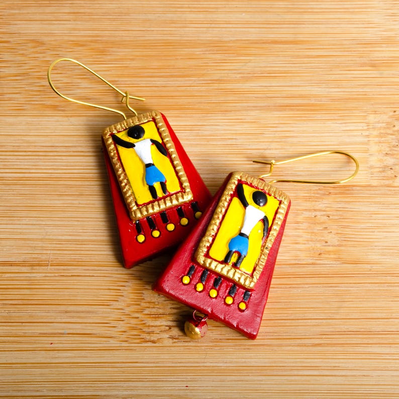May include: A pair of red clay earrings with gold accents. Each earring features a yellow rectangle with a black and white stick figure design and blue accents.