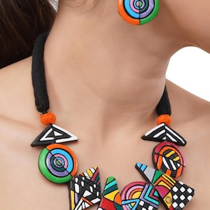 May include: A colorful necklace and earring set featuring geometric shapes in black, white, orange, green, blue, and pink. The necklace has a black cord with orange accents. The earrings are circular with a matching design.