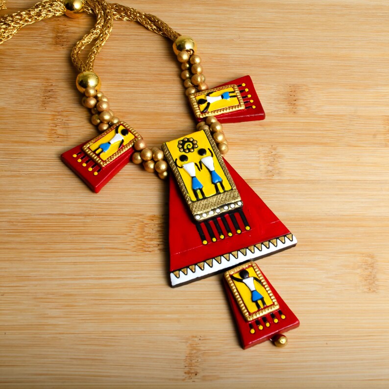 May include: A red, yellow, and blue necklace with a triangular pendant featuring painted figures. The pendant is made of clay and has a gold-colored cord.