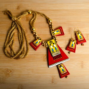 May include: A red, yellow, and blue necklace with a pendant featuring a group of people. The necklace is made of clay and has a gold cord. The pendant is shaped like a triangle and has a white border. The necklace comes with a pair of matching earrings.
