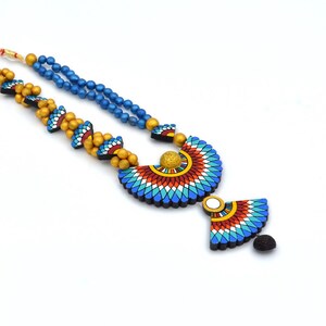 May include: A blue and gold beaded necklace with a large, colorful pendant featuring a geometric design. The pendant is shaped like a crescent moon and has a smaller, matching pendant hanging from it.
