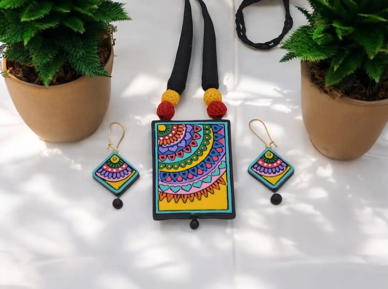 Puede incluir: Un conjunto de collar y pendientes de arcilla de colores. El collar presenta un colgante rectangular con un dise&ntilde;o de mandala azul, verde, amarillo, rosa y morado. Los pendientes son peque&ntilde;os y cuadrados con un dise&ntilde;o de mandala similar.