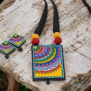 Puede incluir: Un collar de cer&aacute;mica colorido con un colgante cuadrado que presenta un dise&ntilde;o de mandala. El colgante est&aacute; pintado con una variedad de colores, incluyendo amarillo, azul, verde, rosa y morado. El collar est&aacute; acentuado con cord&oacute;n negro y cuentas rojas y amarillas. Tambi&eacute;n se incluyen pendientes a juego con un dise&ntilde;o similar.