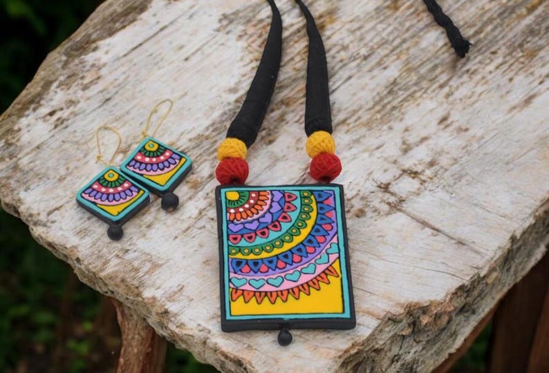 Puede incluir: Un collar de cer&aacute;mica colorido con un colgante cuadrado que presenta un dise&ntilde;o de mandala. El colgante est&aacute; pintado con una variedad de colores, incluyendo amarillo, azul, verde, rosa y morado. El collar est&aacute; acentuado con cord&oacute;n negro y cuentas rojas y amarillas. Tambi&eacute;n se incluyen pendientes a juego con un dise&ntilde;o similar.