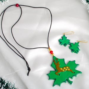 May include: A green maple leaf necklace and earrings set with a gold deer pendant. The necklace has a black cord and red beads. The earrings are gold with green maple leaf charms.