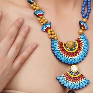 May include: A colorful necklace with a large, blue, red, and yellow pendant in the shape of a stylized wing. The pendant is made of ceramic and has a mosaic-like design. The necklace is strung with gold beads.