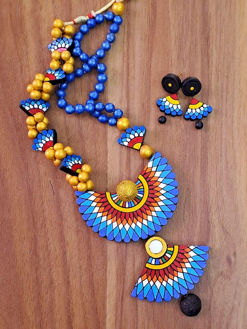 May include: A colorful necklace and earrings set made of terracotta clay. The necklace features a large, geometric pendant with blue, red, yellow, and white patterns. The earrings are smaller versions of the pendant, with black clay accents.