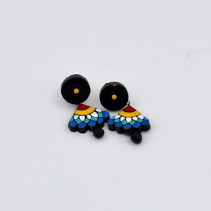 May include: A pair of black clay earrings with a colorful geometric design. The earrings feature a black circle with a yellow dot at the top and a triangular pendant with blue, white, yellow, and red details.