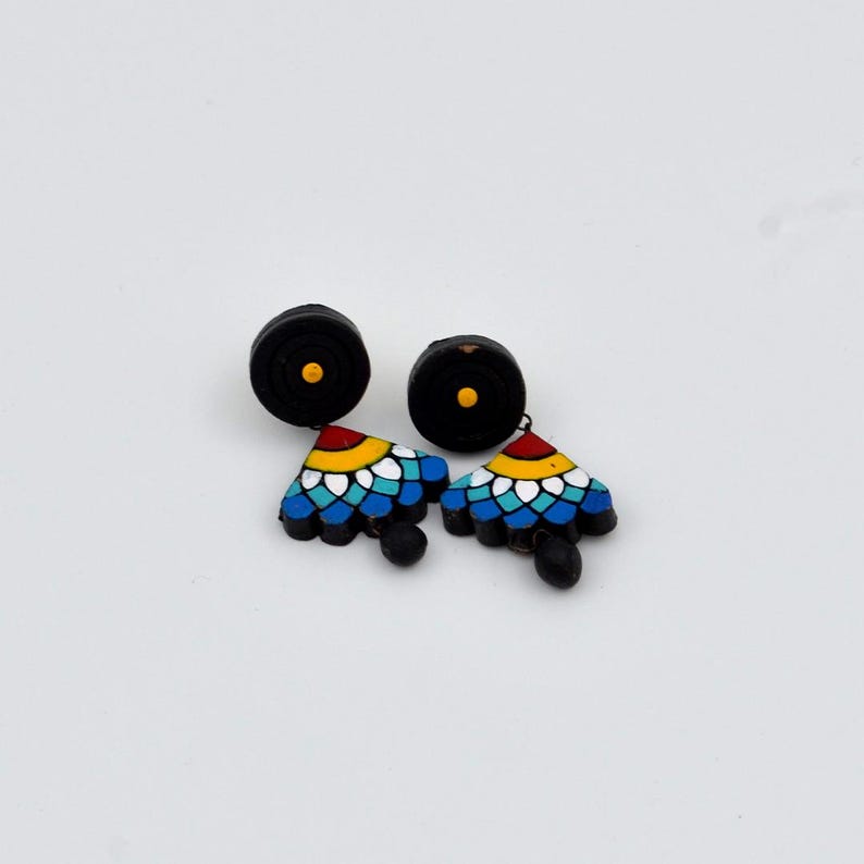May include: A pair of black clay earrings with a colorful geometric design. The earrings feature a black circle with a yellow dot at the top and a triangular pendant with blue, white, yellow, and red details.