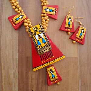 May include: A red and gold necklace with a large triangular pendant featuring a painted design of two figures. The pendant is decorated with black, yellow, and blue accents. The necklace is paired with matching earrings with a similar design.