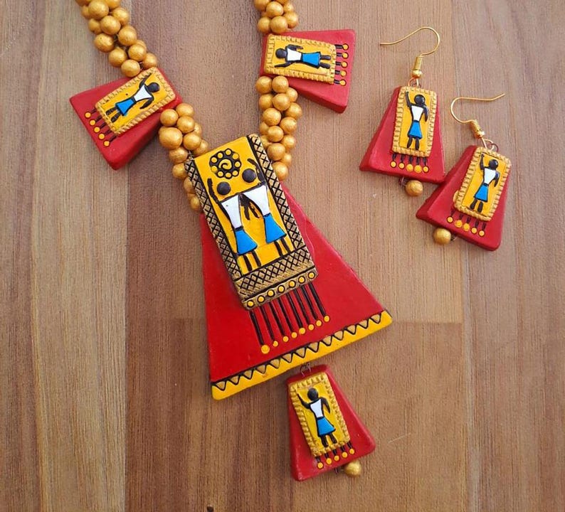 May include: A red and gold necklace with a large triangular pendant featuring a painted design of two figures. The pendant is decorated with black, yellow, and blue accents. The necklace is paired with matching earrings with a similar design.