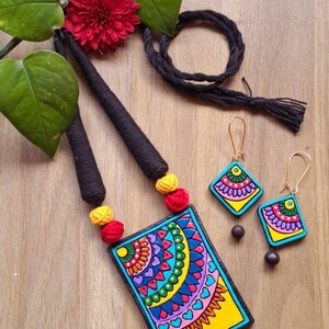 Puede incluir: Un conjunto de collar y pendientes de terracota coloridos con un dise&ntilde;o geom&eacute;trico. El collar presenta un colgante rectangular con un fondo amarillo y un patr&oacute;n de mandala multicolor. Los pendientes son de forma cuadrada con un dise&ntilde;o de mandala similar.