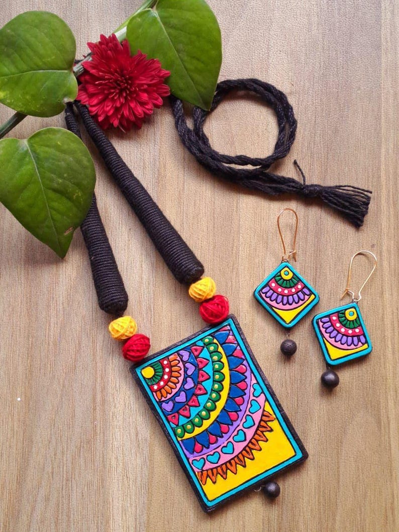 Puede incluir: Un conjunto de collar y pendientes de terracota coloridos con un dise&ntilde;o geom&eacute;trico. El collar presenta un colgante rectangular con un fondo amarillo y un patr&oacute;n de mandala multicolor. Los pendientes son de forma cuadrada con un dise&ntilde;o de mandala similar.