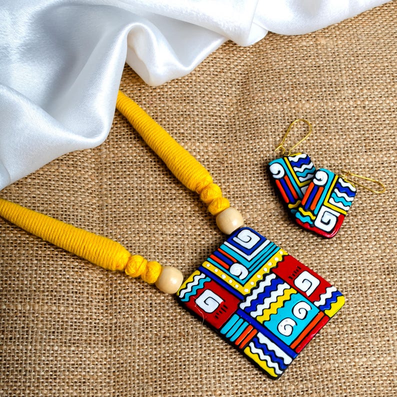 Puede incluir: Un conjunto de collar y pendientes hechos a mano. El collar presenta un cord&oacute;n amarillo con un colgante geom&eacute;trico en rojo, azul, amarillo y blanco. Los pendientes a juego tienen un dise&ntilde;o similar. El conjunto de joyas se muestra sobre una superficie texturizada.