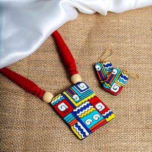 Puede incluir: Un collar y un juego de pendientes a juego. El colgante y los pendientes presentan un dise&ntilde;o geom&eacute;trico con detalles en rojo, amarillo, azul y blanco. El collar tiene un cord&oacute;n rojo y cuentas de madera. Los pendientes tienen ganchos dorados.