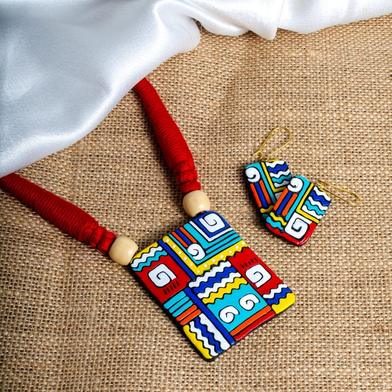 Puede incluir: Un collar y un juego de pendientes a juego. El colgante y los pendientes presentan un dise&ntilde;o geom&eacute;trico con detalles en rojo, amarillo, azul y blanco. El collar tiene un cord&oacute;n rojo y cuentas de madera. Los pendientes tienen ganchos dorados.