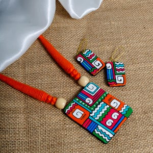 Puede incluir: Un vibrante conjunto de collar y pendientes con un dise&ntilde;o geom&eacute;trico. El collar tiene un cord&oacute;n naranja y un colgante cuadrado grande con un patr&oacute;n colorido de formas rojas, verdes, azules y blancas. Los pendientes a juego son rectangulares.