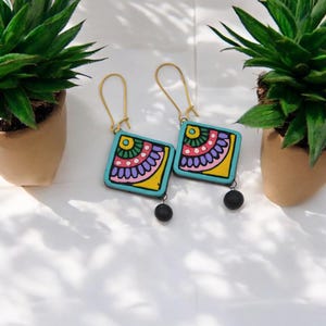 Puede incluir: Un par de pendientes cuadrados de color azul turquesa con dise&ntilde;os florales coloridos y detalles de cuentas negras. Los pendientes tienen ganchos dorados.